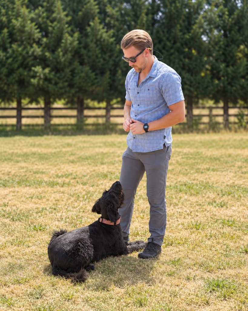 Cameron standing beside his black dog lying on a grassy training field.